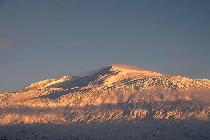 A wide, snow-covered mountain glowing in the warm light of sunrise or sunset.